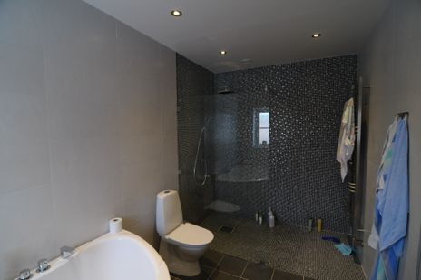 Badrum med vita väggar, spotlights i taket, vit wc-stol, mosaikvägg i grått