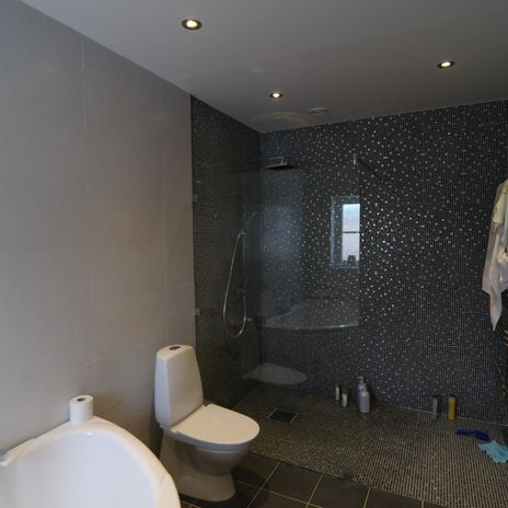 Badrum med vita väggar, spotlights i taket, vit wc-stol, mosaikvägg i grått