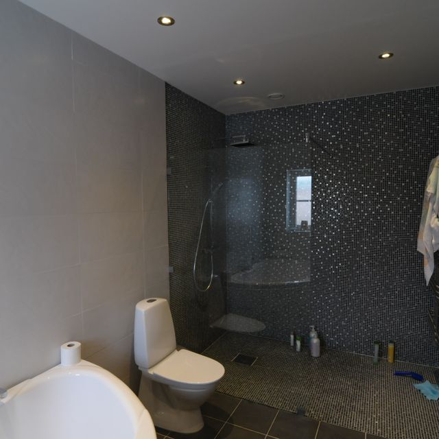 Badrum med vita väggar, spotlights i taket, vit wc-stol, mosaikvägg i grått
