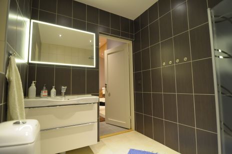 Badrum med grå kakelplattor på väggen, vit handfatskomod, badrumsspegle med belysning. 