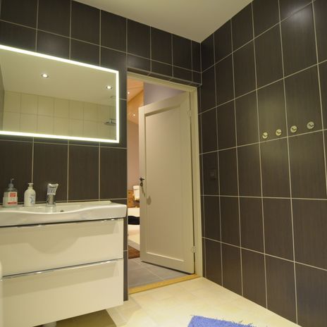 Badrum med grå kakelplattor på väggen, vit handfatskomod, badrumsspegle med belysning. 