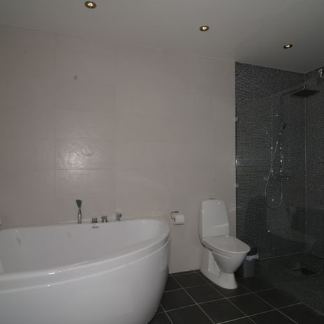 Vitt badrum med vitt hörnbadkar, vit wc-stol, glasduschvägg